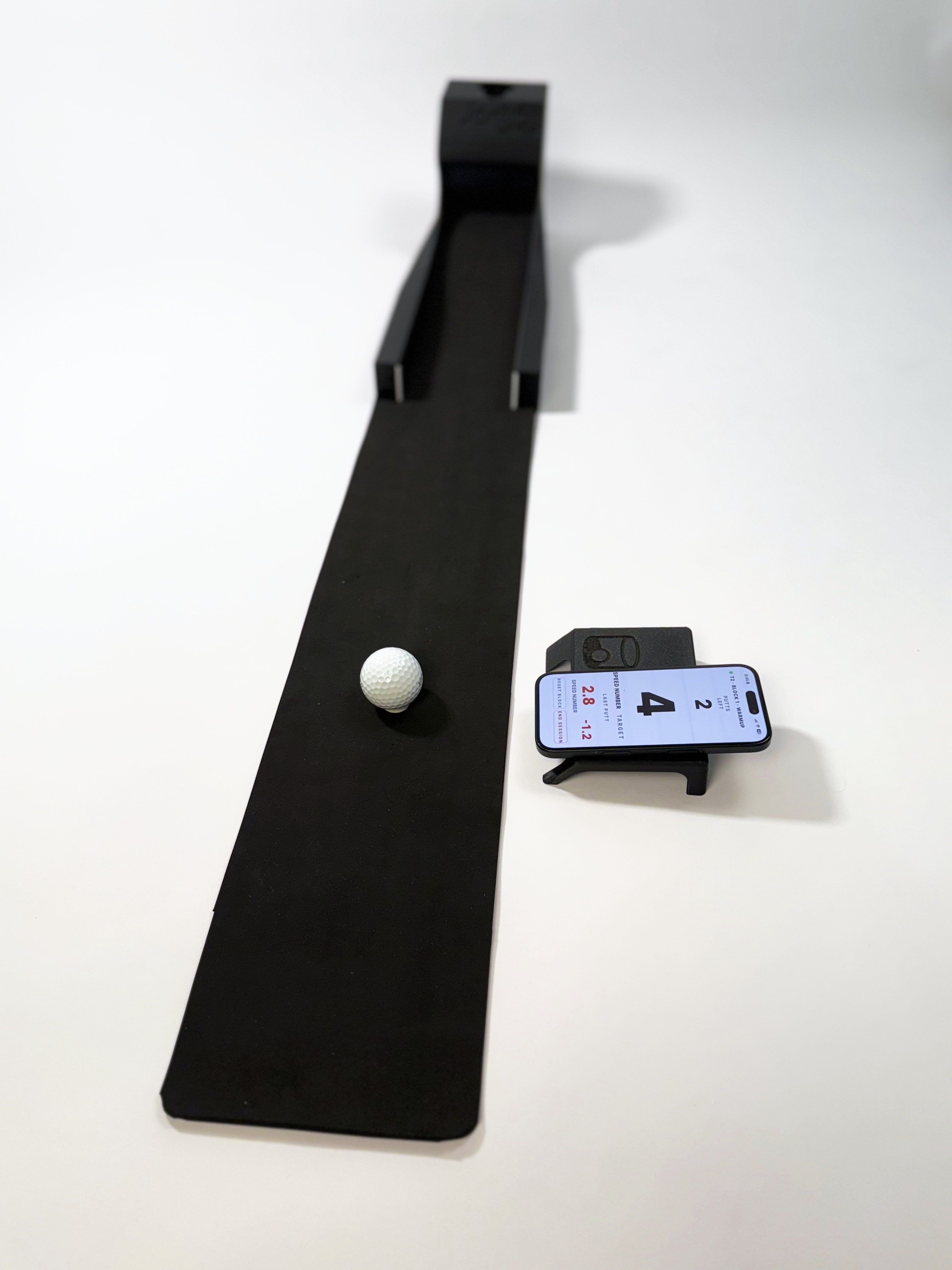 The Speed Machine from the hitting end: the full length of putting mat running up to the ball-return tower, a ball on the mat and an iPhone on its stand showing the Jarit App